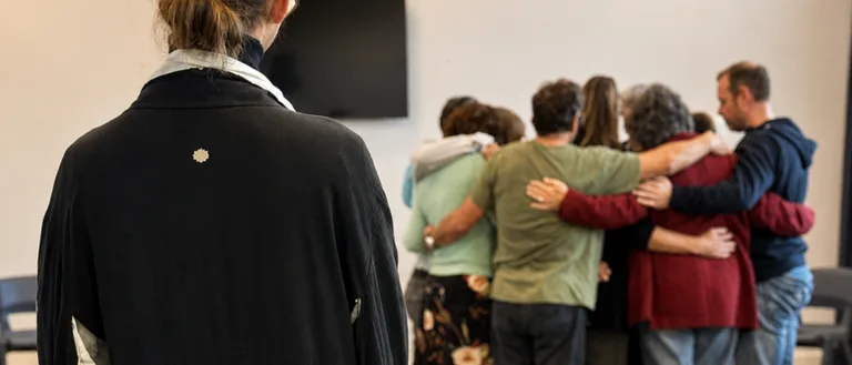 Begeleider kijkt toe terwijl de groep na een familieopstelling in een omhelzing bij elkaar staat